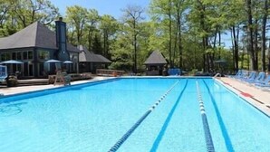 A swimming pool with clear lanes, surrounded by lounge chairs and shaded areas, with trees in the background.