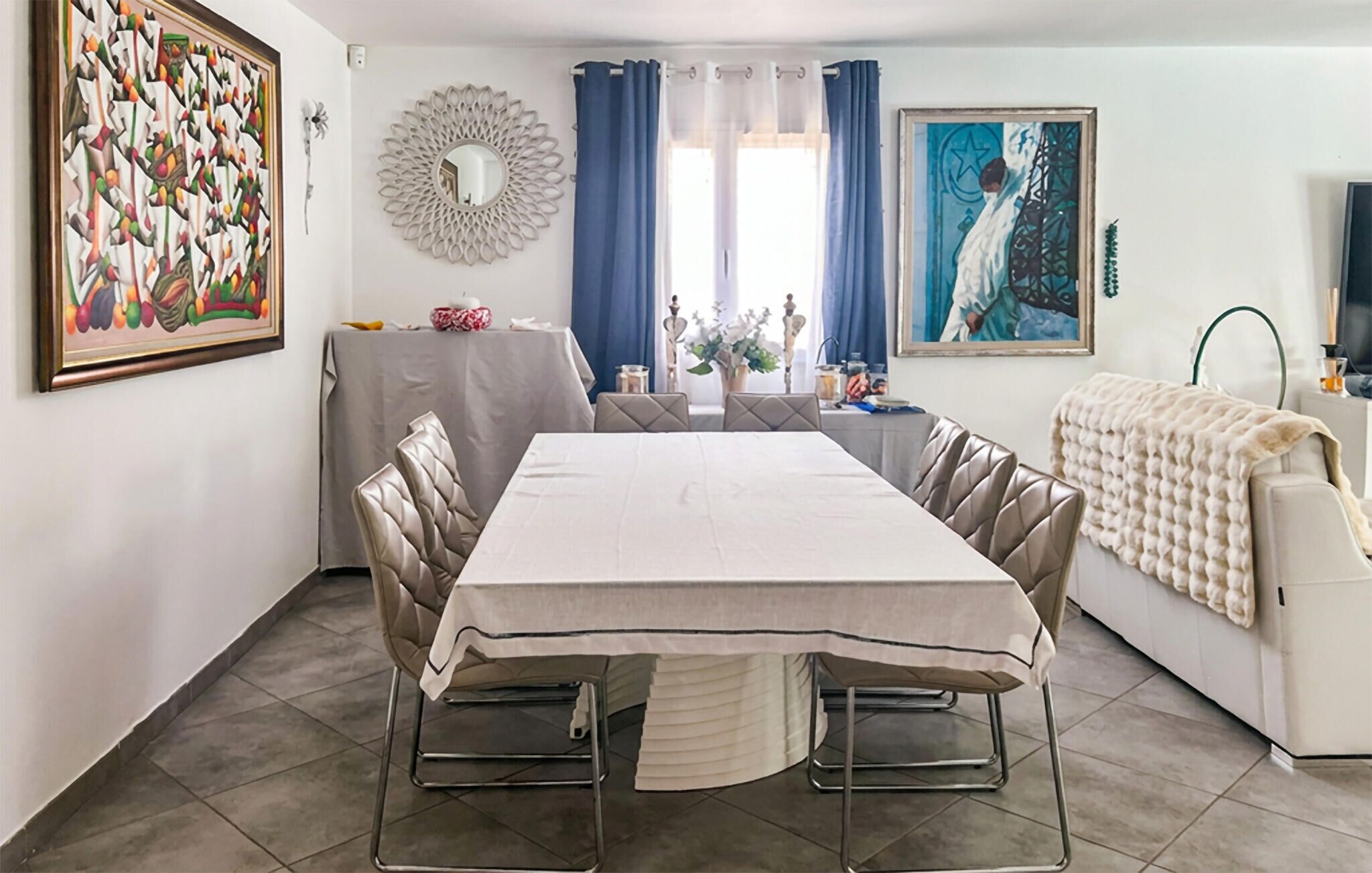 Un comedor con una mesa larga, sillas con cojines, un espejo decorativo y obras de arte en las paredes.