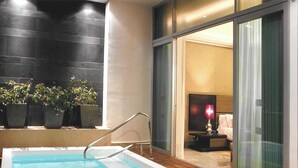 Una piscina interior moderna con una terraza de madera, plantas en macetas y vistas a un dormitorio a través de puertas de cristal.