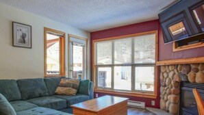 A living room with a sectional sofa, a wooden coffee table, a stone fireplace, and large windows with blinds. - Fantastic location; 2 bdrm w/ private hot tub (Sun Peaks)