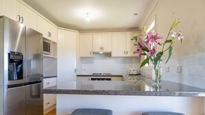 A modern kitchen with stainless steel appliances, white cabinetry, and a central island with bar stools. - Vineyard Cottage - Your Innkeeper Mudgee (Eurunderee)