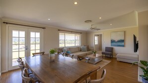 A spacious living area with a wooden dining table, sofas, and a large window with blinds. - Vineyard Cottage - Your Innkeeper Mudgee (Eurunderee)