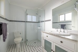 A bathroom with a glass-enclosed shower, a white toilet, a sink with a mirror, and a tiled floor. - Vineyard Cottage - Your Innkeeper Mudgee (Eurunderee)
