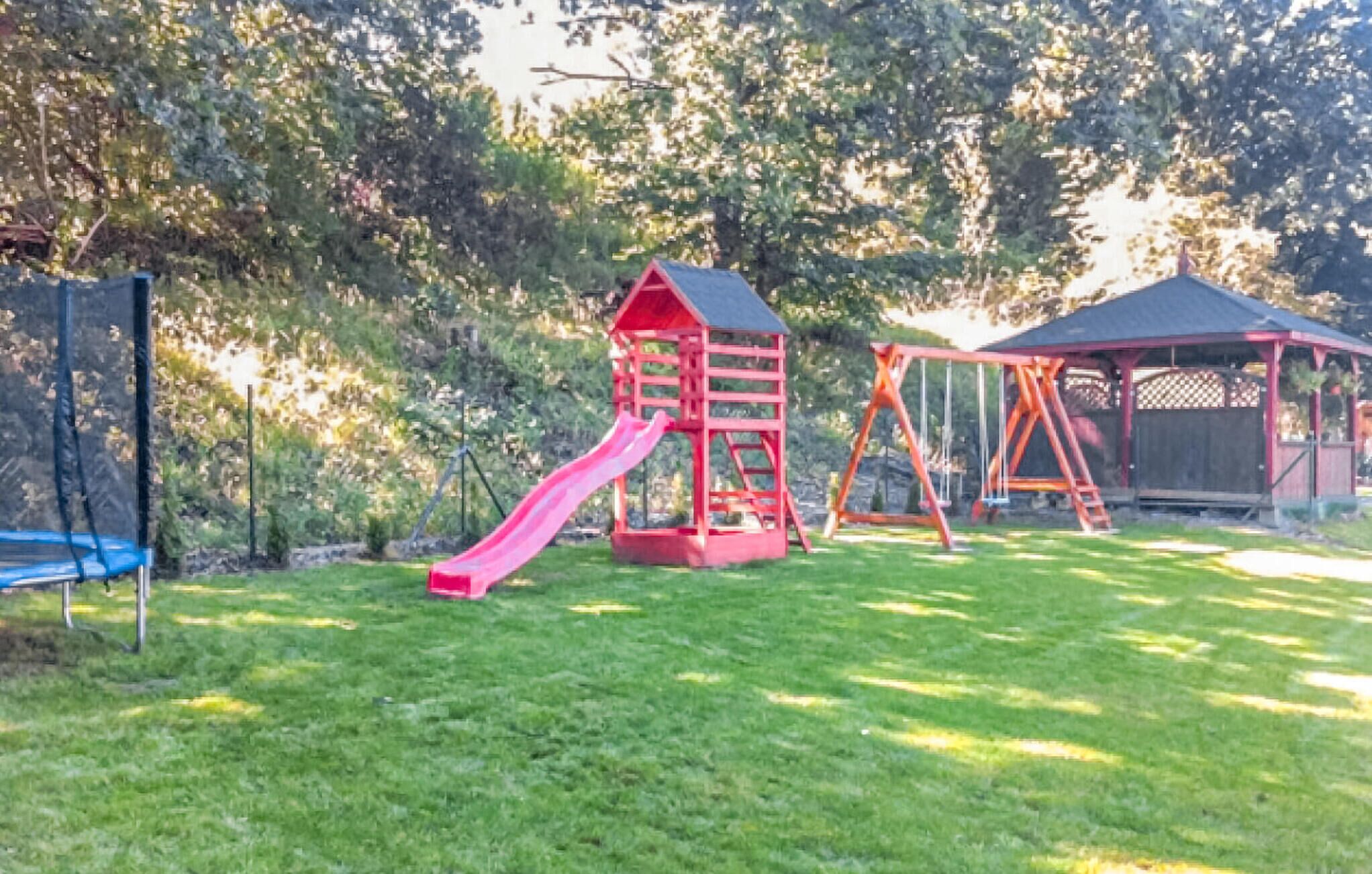 A playground with a red slide and swings, surrounded by trees and a fenced area.