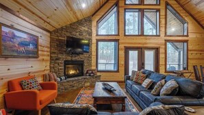 A cozy cabin interior with a stone fireplace, wooden walls, a large window, and a comfortable seating area. - Over Yonder-3Bed/Bunks/Pool Table/3.5 Bath (Broken Bow)