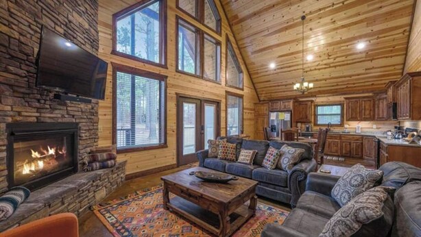 A spacious living room with a stone fireplace, large windows, a wooden ceiling, and a cozy seating area with a sofa and armchairs. - Over Yonder-3Bed/Bunks/Pool Table/3.5 Bath (Broken Bow)