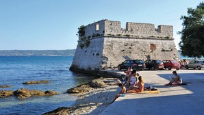 Una zona costera con una fortificación de piedra, una playa con gente tomando el sol y coches aparcados a lo largo de la orilla.