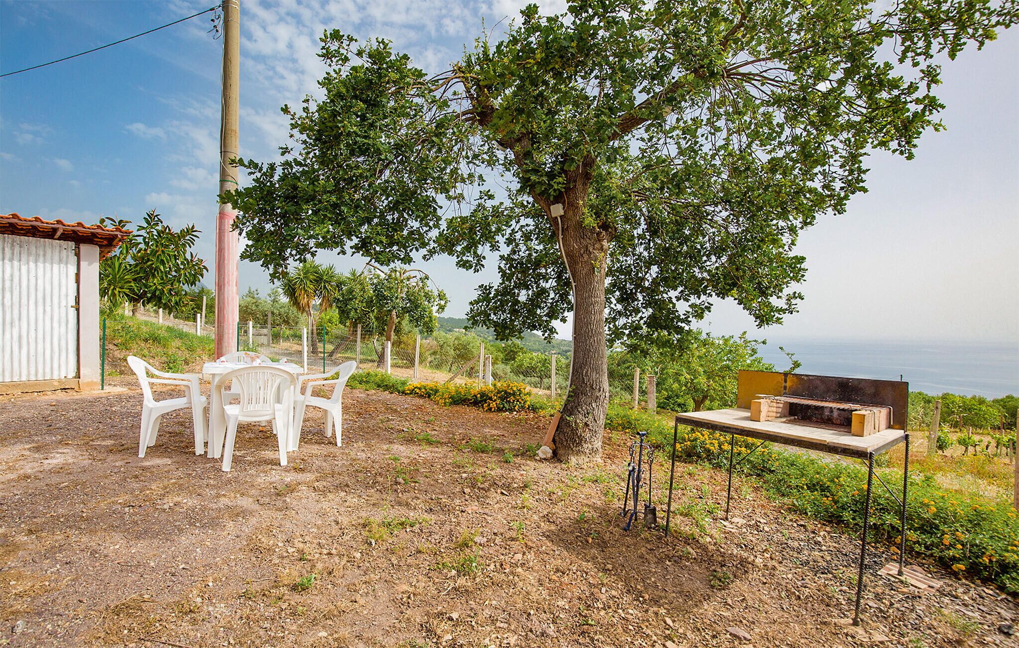 A rustic outdoor seating area with a table and chairs, a barbecue grill, and a view of the sea.