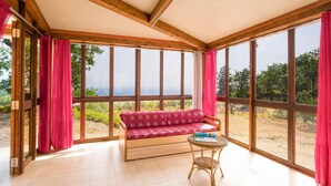 Une pièce avec un plafond en bois, de grandes fenêtres, un canapé rouge à motifs et une table en osier.