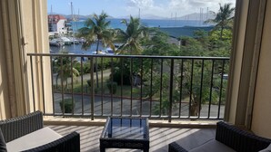 Een balkon met rieten meubilair, uitzicht op een jachthaven met boten, palmbomen en verre bergen.