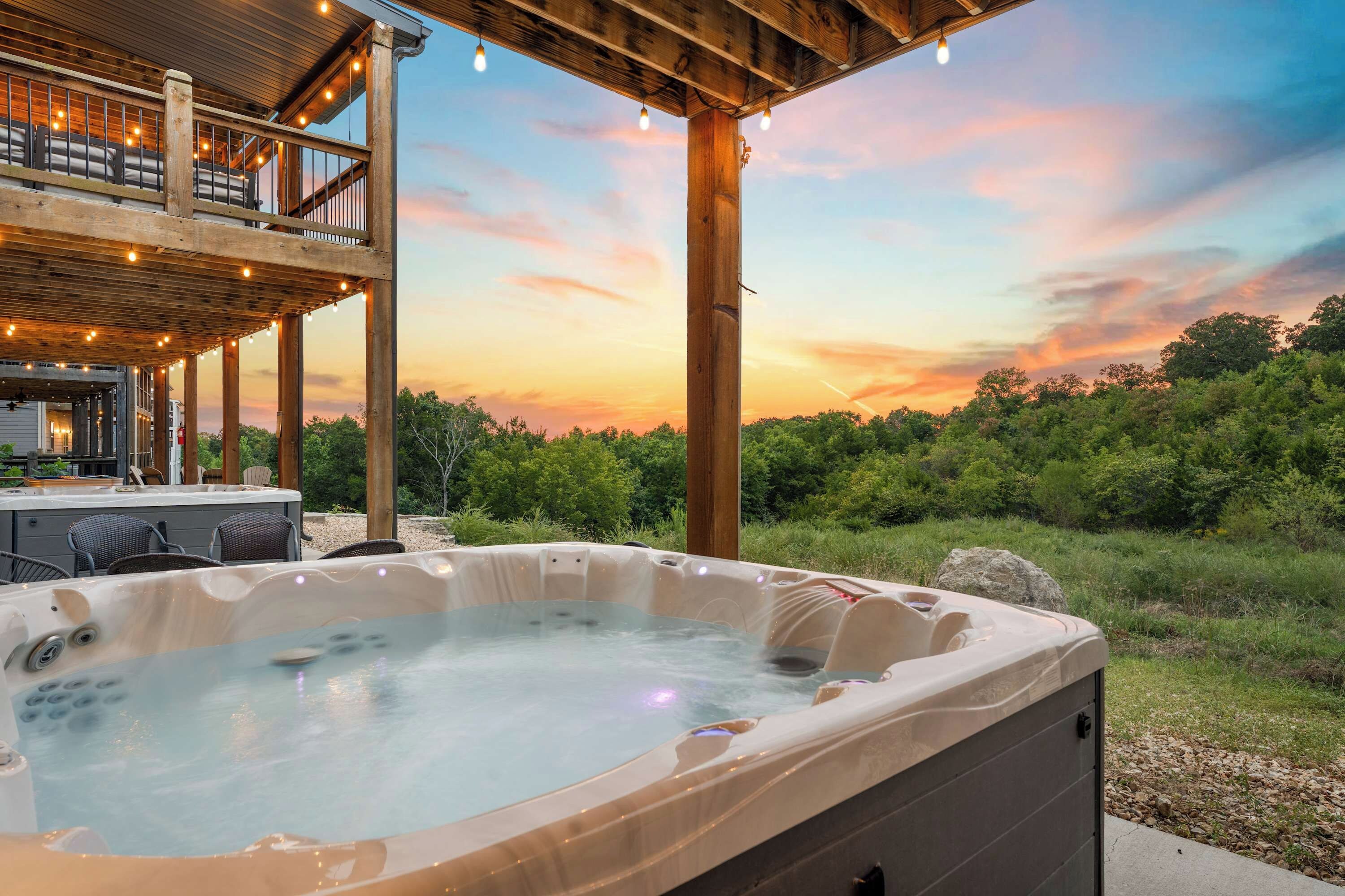A hot tub with a wooden deck and lounge chairs, surrounded by greenery and a sunset view.