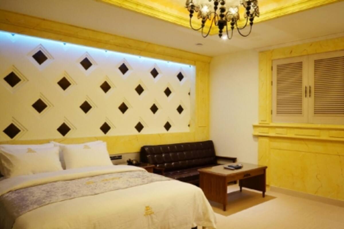 A bedroom with a bed, a sofa, a small table, a decorative wall panel, and a window with shutters.