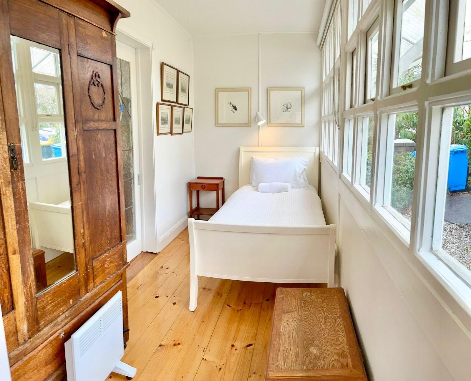 A bedroom with a wooden floor, a white bed, a small bedside table, a wooden bench, and a large window with a view of greenery.