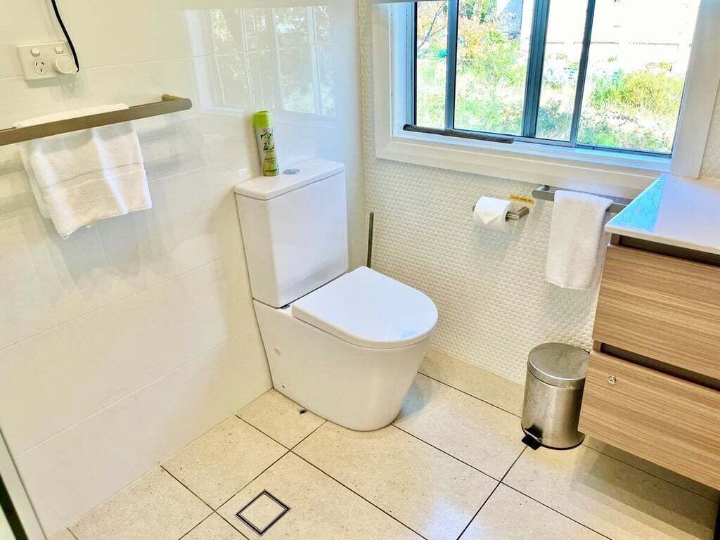A small bathroom with a toilet, a trash bin, a towel rack, and a window with a view of greenery outside.