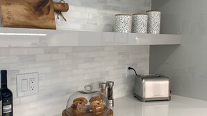 A kitchen with white subway tiles, a wooden cutting board, a glass display case with muffins, a toaster, and a wine bottle. - Casa del Mare - Beautiful Naples Pool Home (Naples)