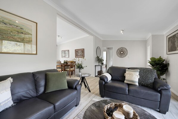 A modern living room with two black leather sofas, a coffee table, and a dining area in the background.