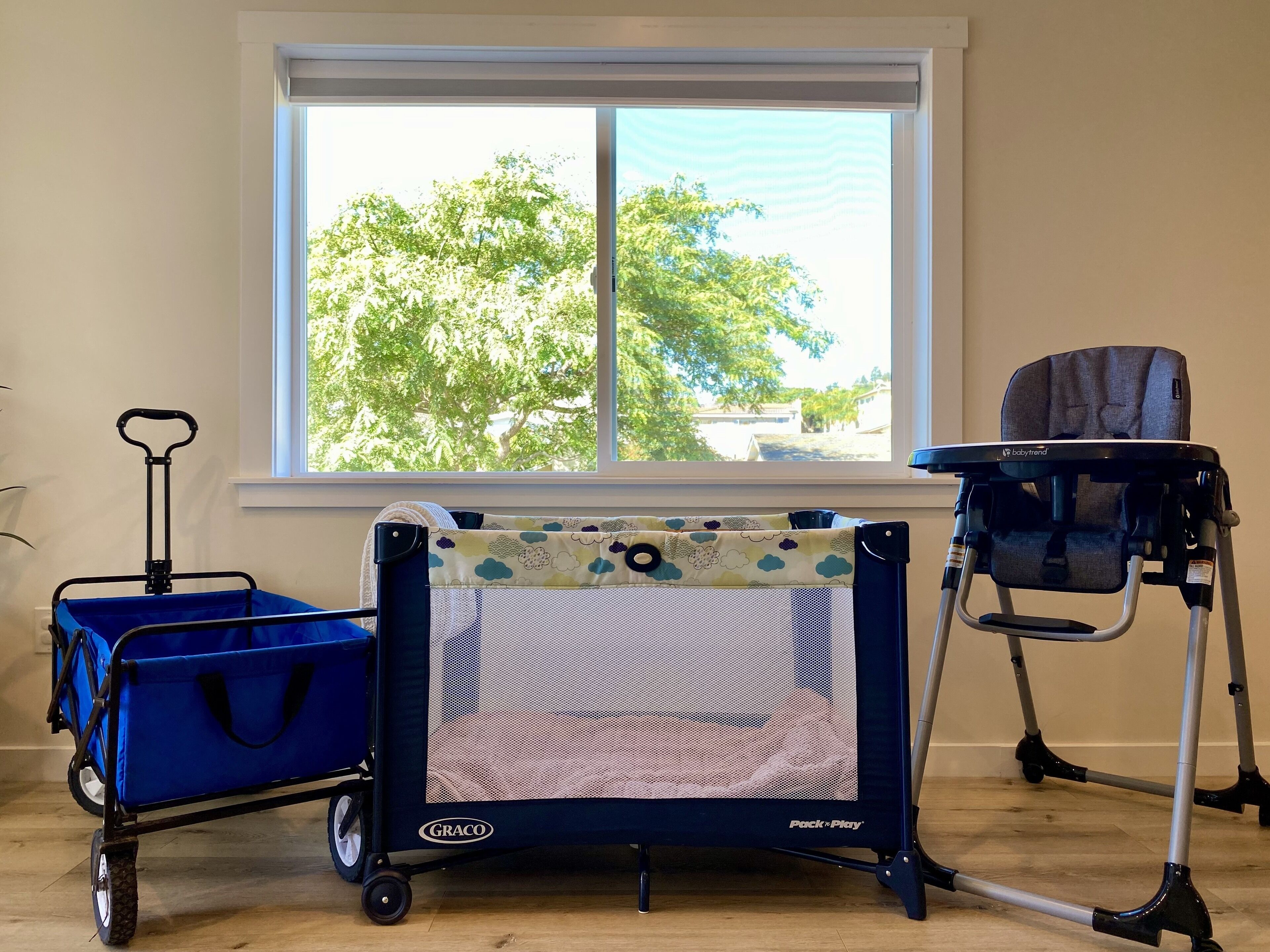 Ein Zimmer mit Fenster, einem blauen Kinderwagen, einem Babyreisebett und einem Kinderwagenkorb.
