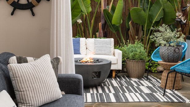 Un salon accueillant avec un canapé gris, une table basse ronde et un coussin à rayures. Un décor représentant un gouvernail de navire est accroché au mur, et de grandes plantes vertes se trouvent à l’arrière-plan.