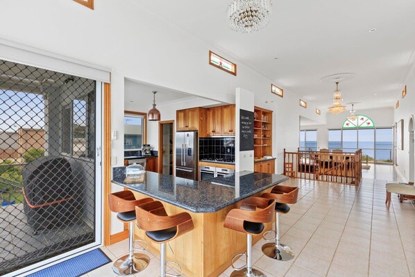 A modern kitchen with a central island, wooden bar stools, and a view of the sea.