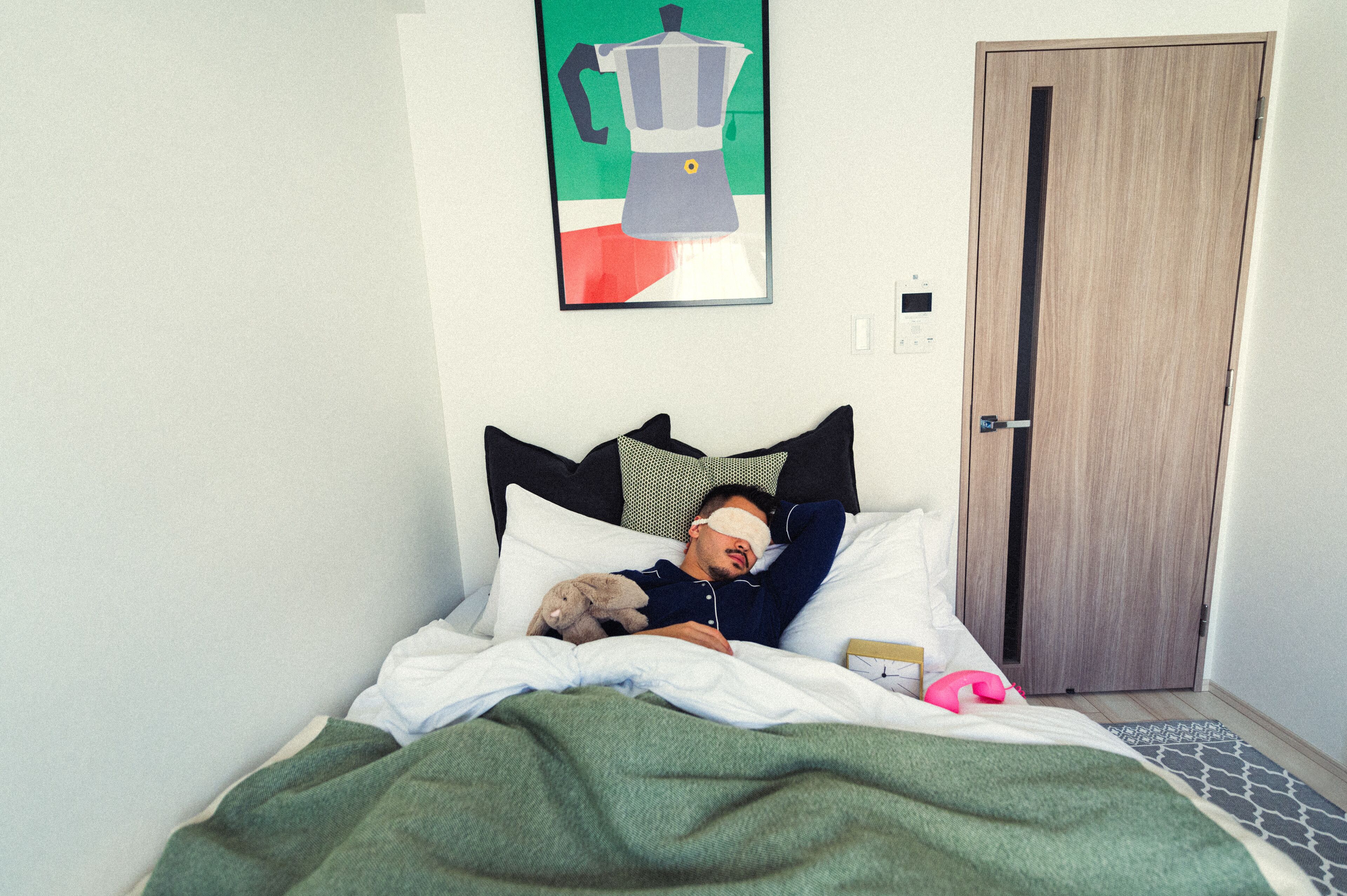 A bed with a green blanket, white pillows, and a teddy bear. There is a door and a wall with a framed picture.