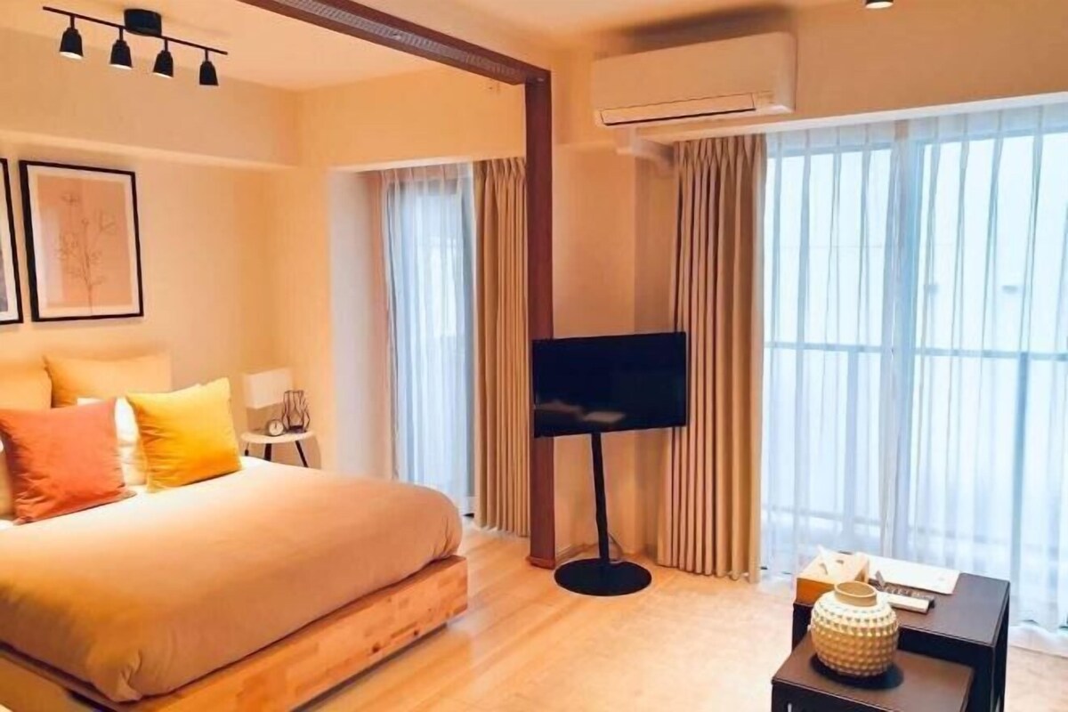 A hotel room with a bed, a flat-screen TV, a side table, and a large window with curtains.