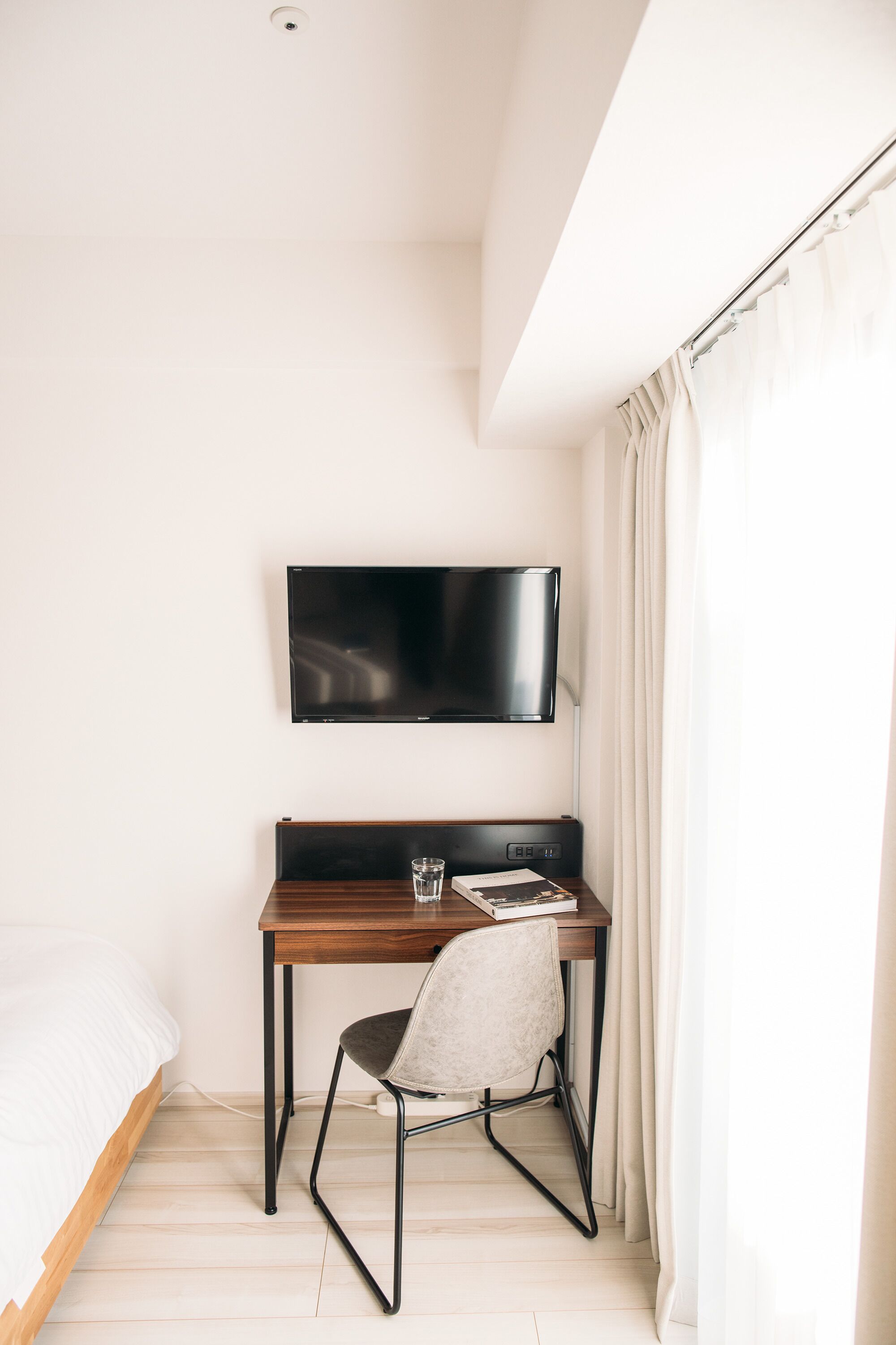 A small, modern room with a bed, a desk with a chair, a flat-screen TV, and a window with curtains.