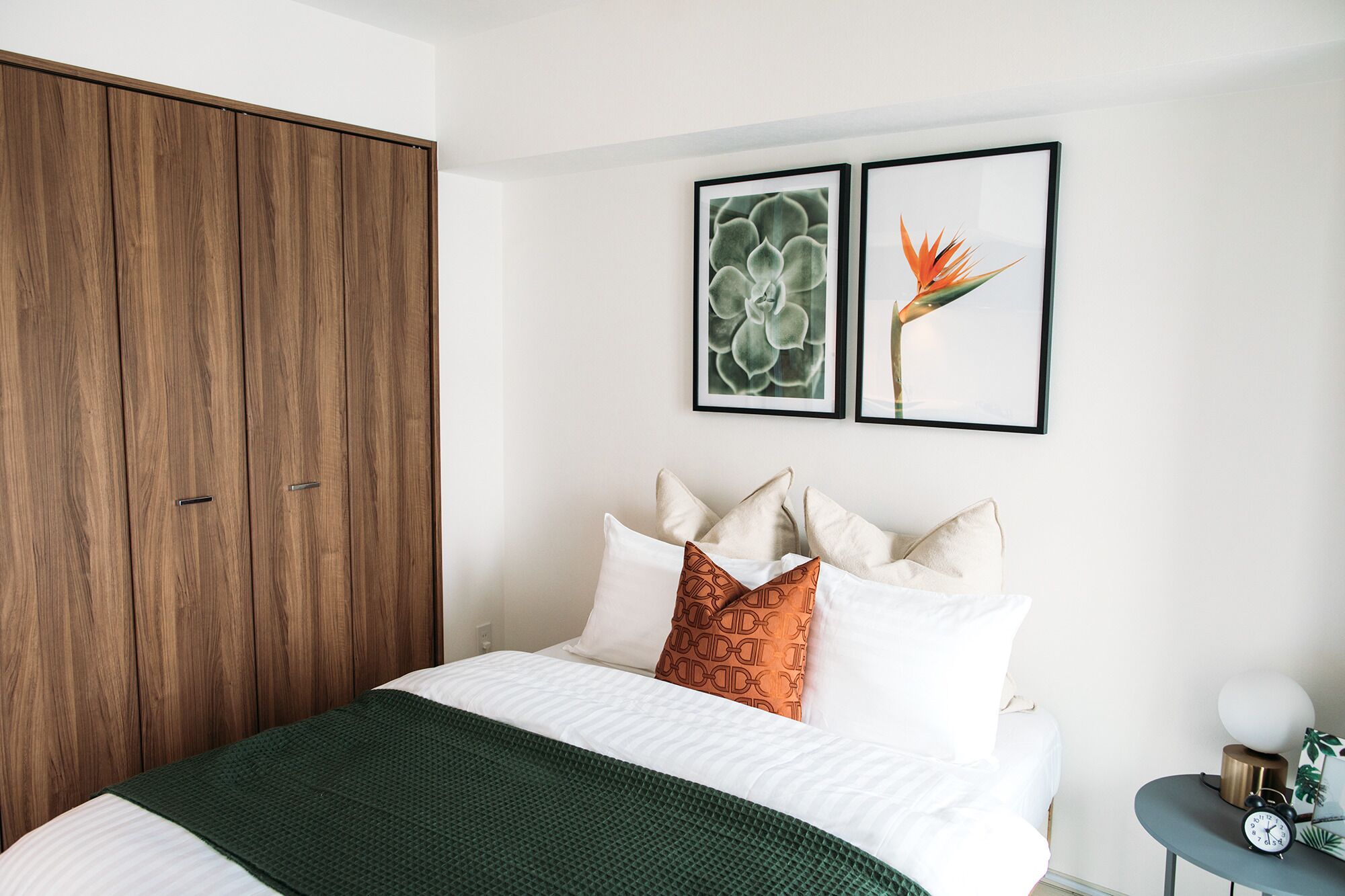 Un dormitorio con una cama, almohadas, una manta verde, un armario de madera y dos cuadros en la pared.