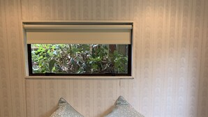 Cama com roupa de cama estampada e várias almofadas, janela com vista para o verde e parede com padrão discreto.