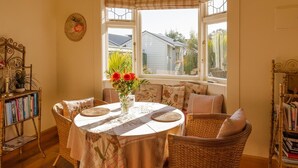 A sunlit room with a round table, wicker chairs, a bookshelf, and a window with a view of a house.