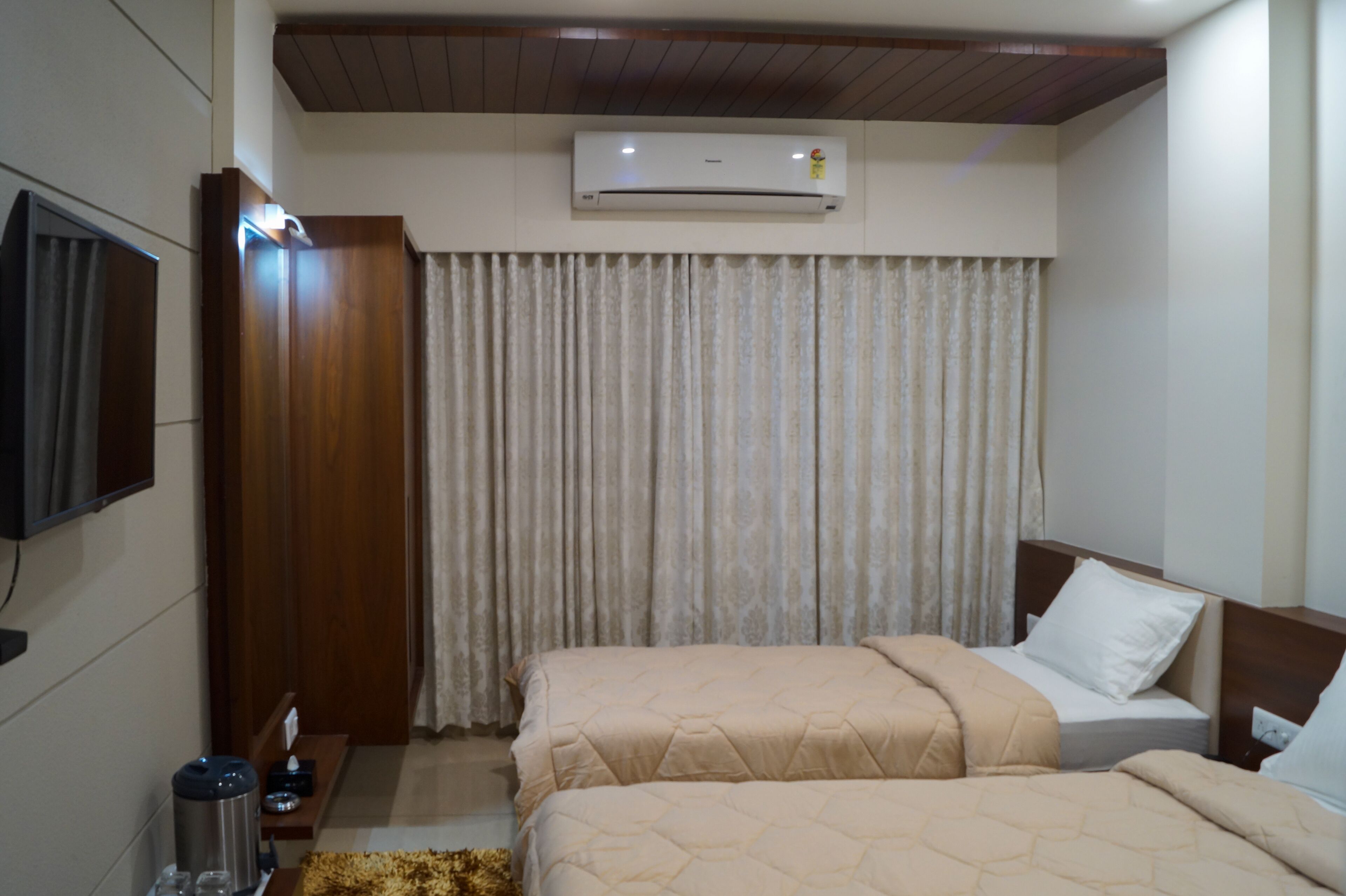 A hotel room with two beds, a TV mounted on the wall, an air conditioning unit, and a wooden ceiling.