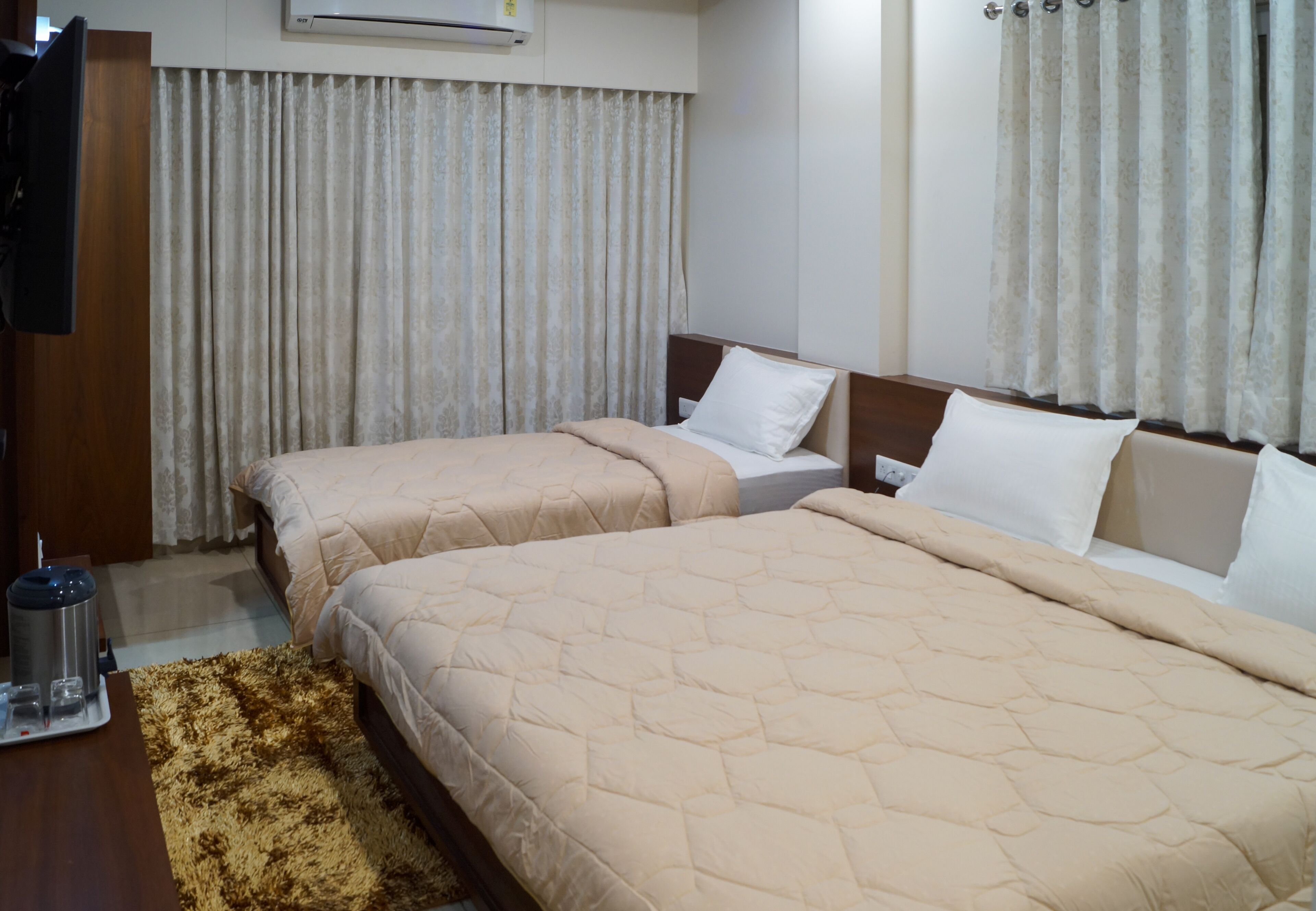 A hotel room with two beds, a wooden wardrobe, a flat-screen TV, and a small table with a tray of glasses and a thermos.