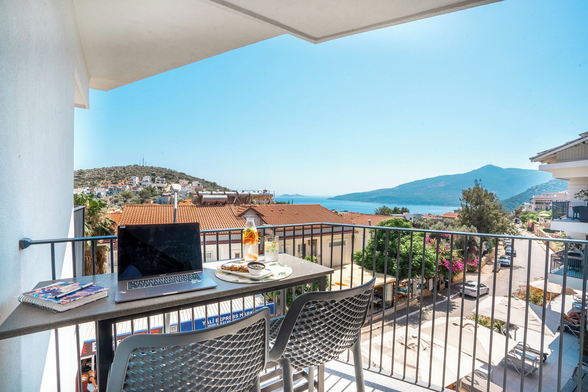 A balcony with a laptop, notebook, and a drink, overlooking a coastal town with buildings and a mountain in the distance.