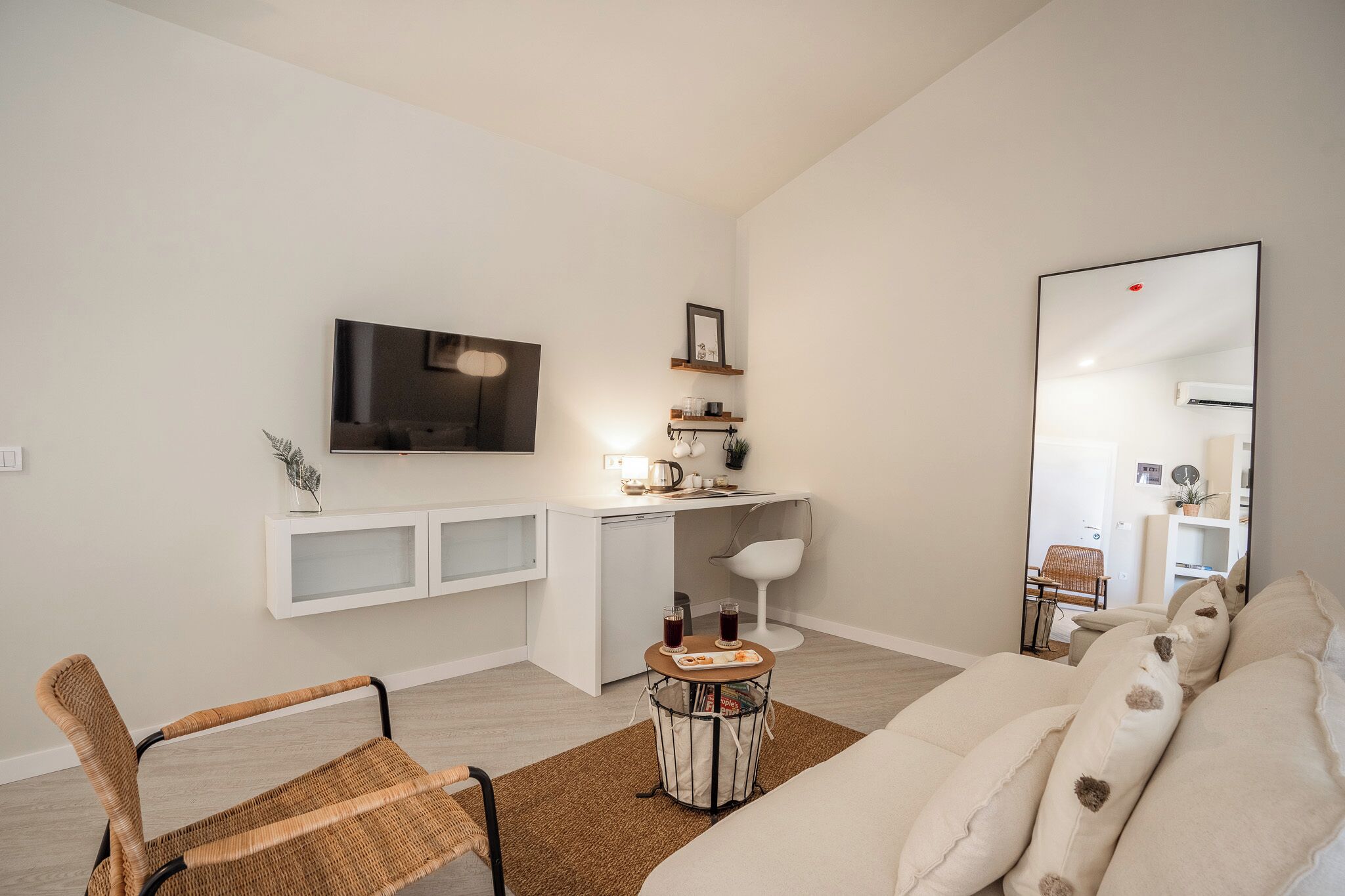Una sala de estar moderna con un televisor de pantalla plana, un escritorio blanco, una silla blanca, una silla de mimbre, una mesita redonda con bebidas y un espejo grande.