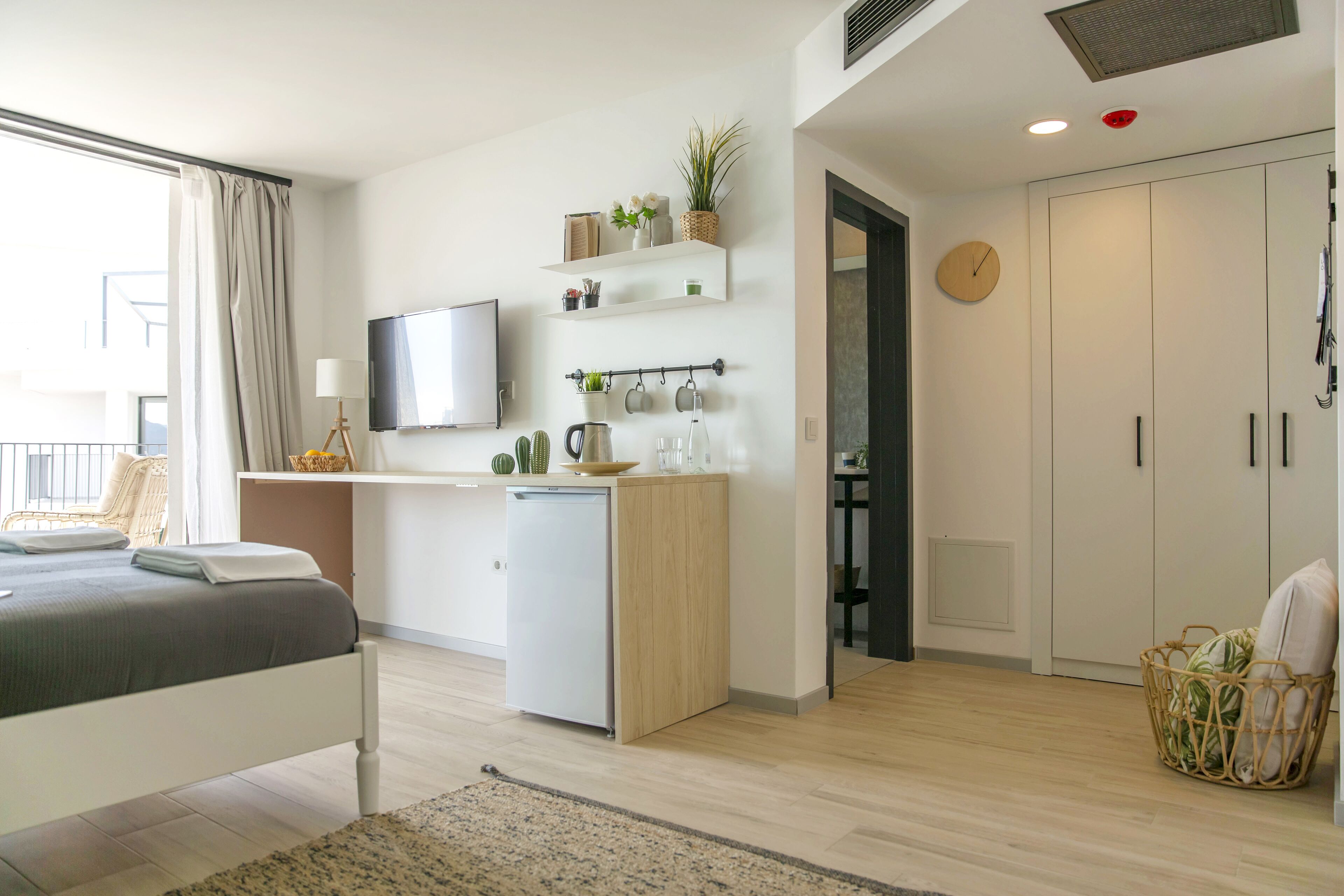 A modern hotel room with a bed, a small kitchenette, a TV, and a wardrobe.