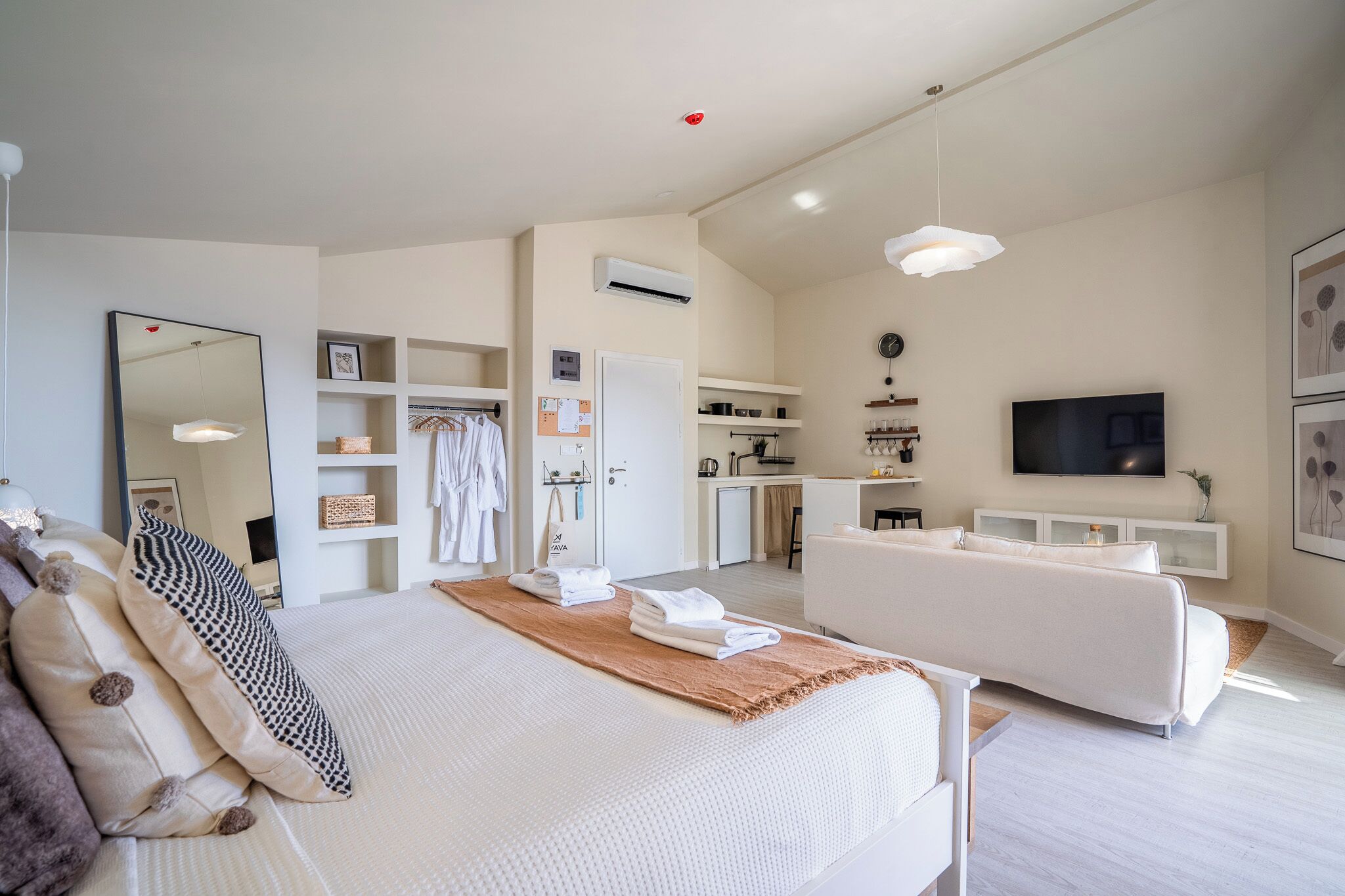 A modern hotel room with a large bed, a sofa, a TV, and a kitchenette.