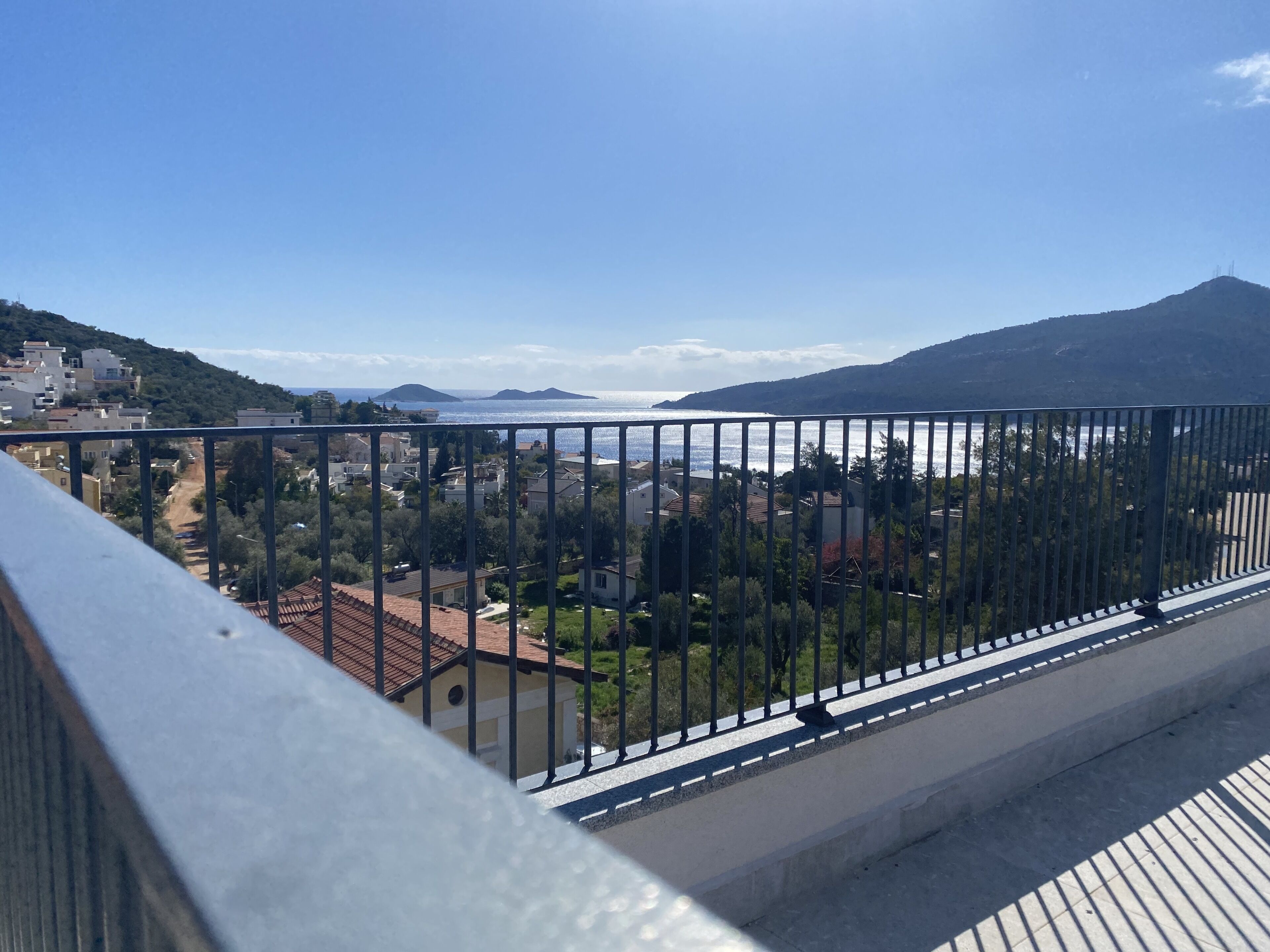 A balcony with a metal railing overlooking a coastal town with buildings and hills in the background.