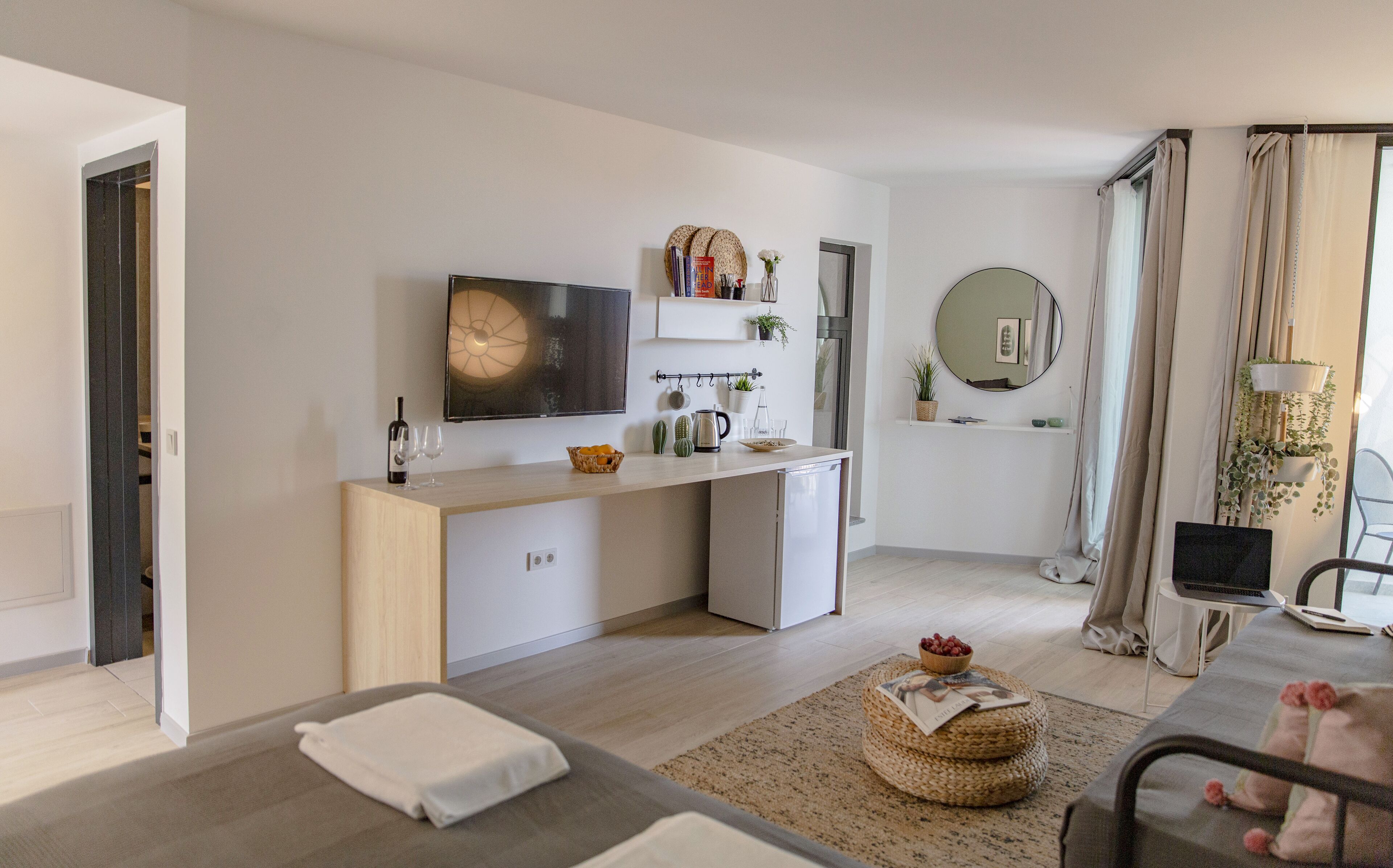 A modern, well-lit room with a minimalist design, featuring a central kitchen area, a dining table with a laptop, and a cozy seating area with a rug and ottoman.