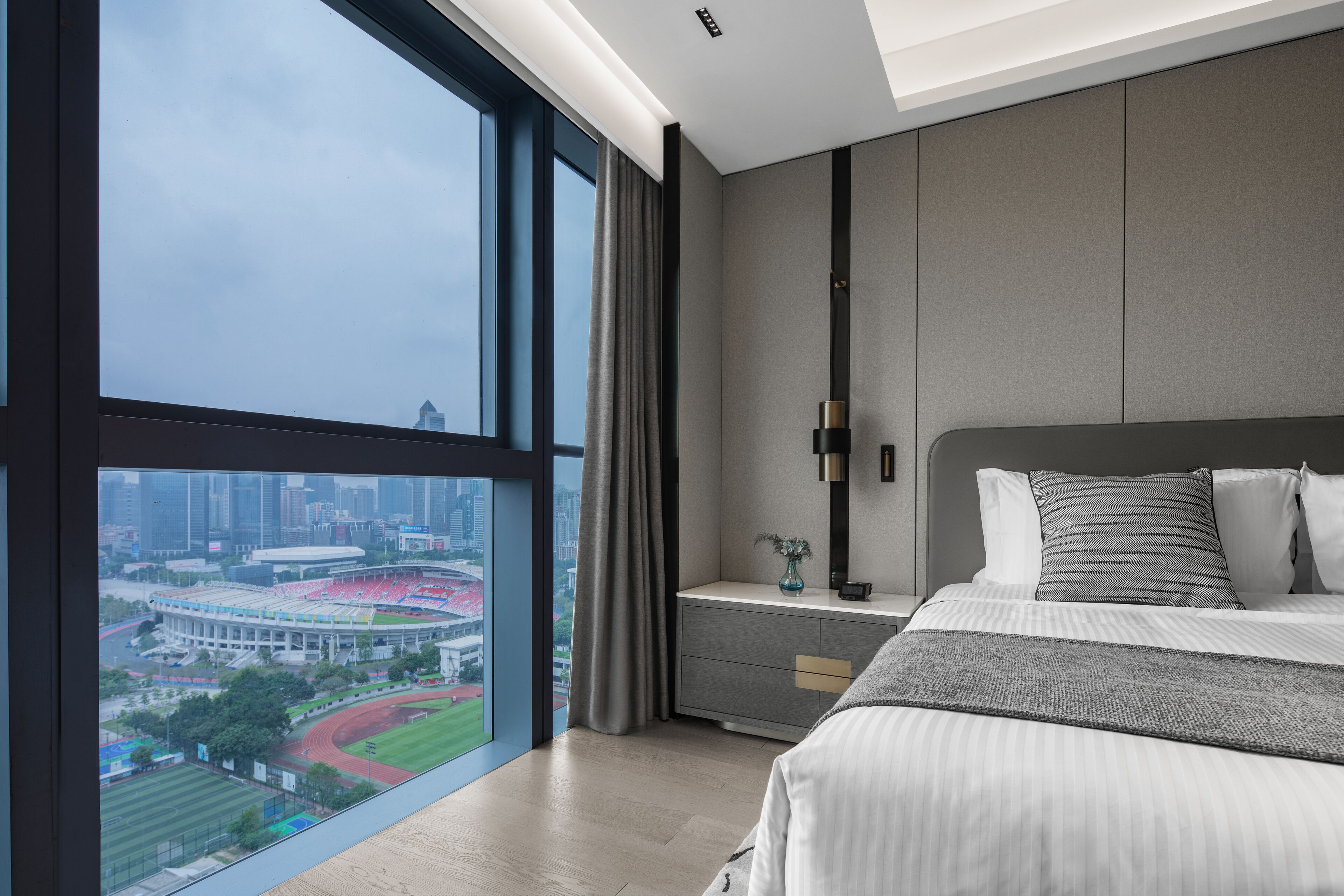 A modern hotel room with a large window offering a city view, a neatly made bed, and a minimalist nightstand.