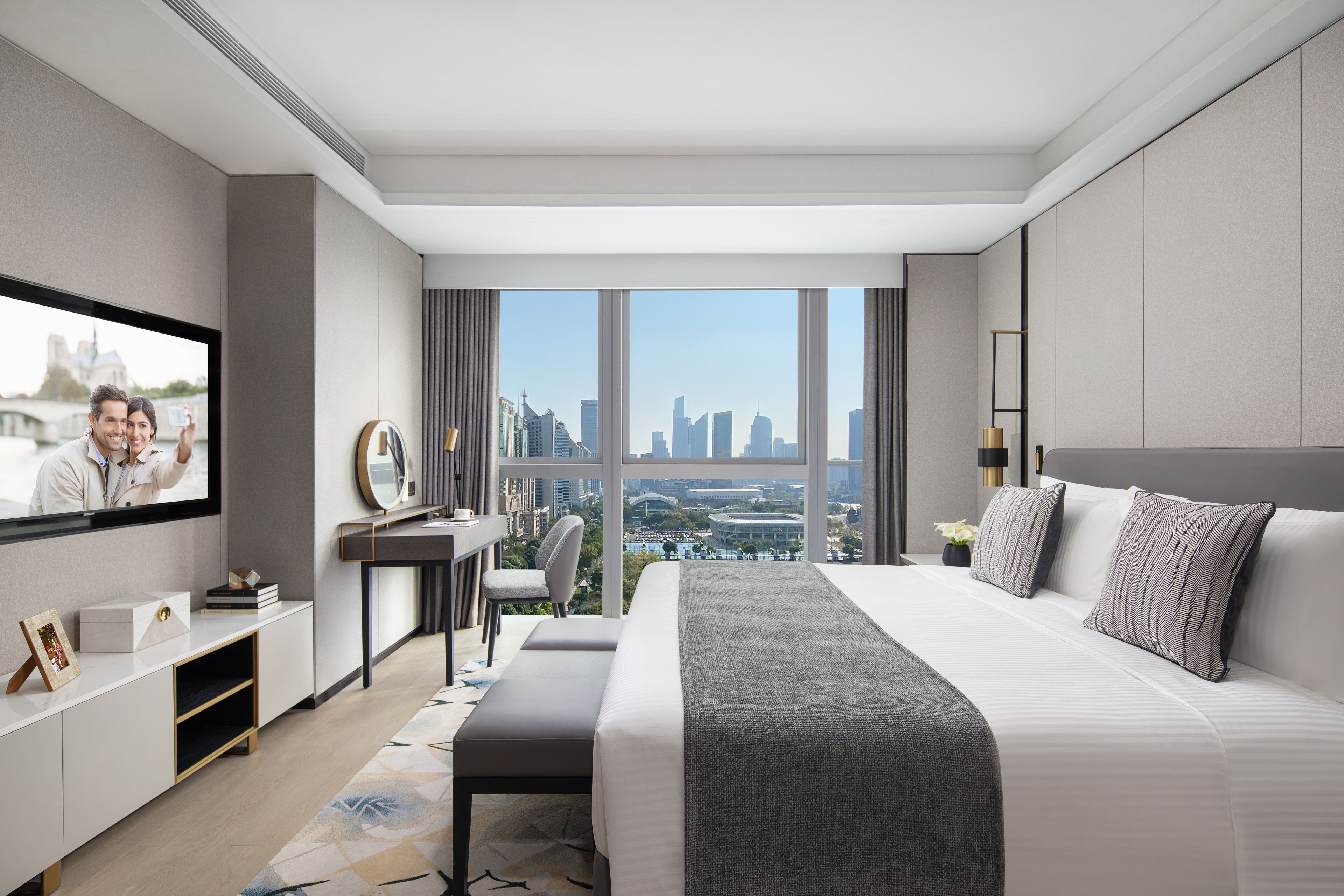 A modern hotel room with a large bed, a desk, a TV, and a city view.