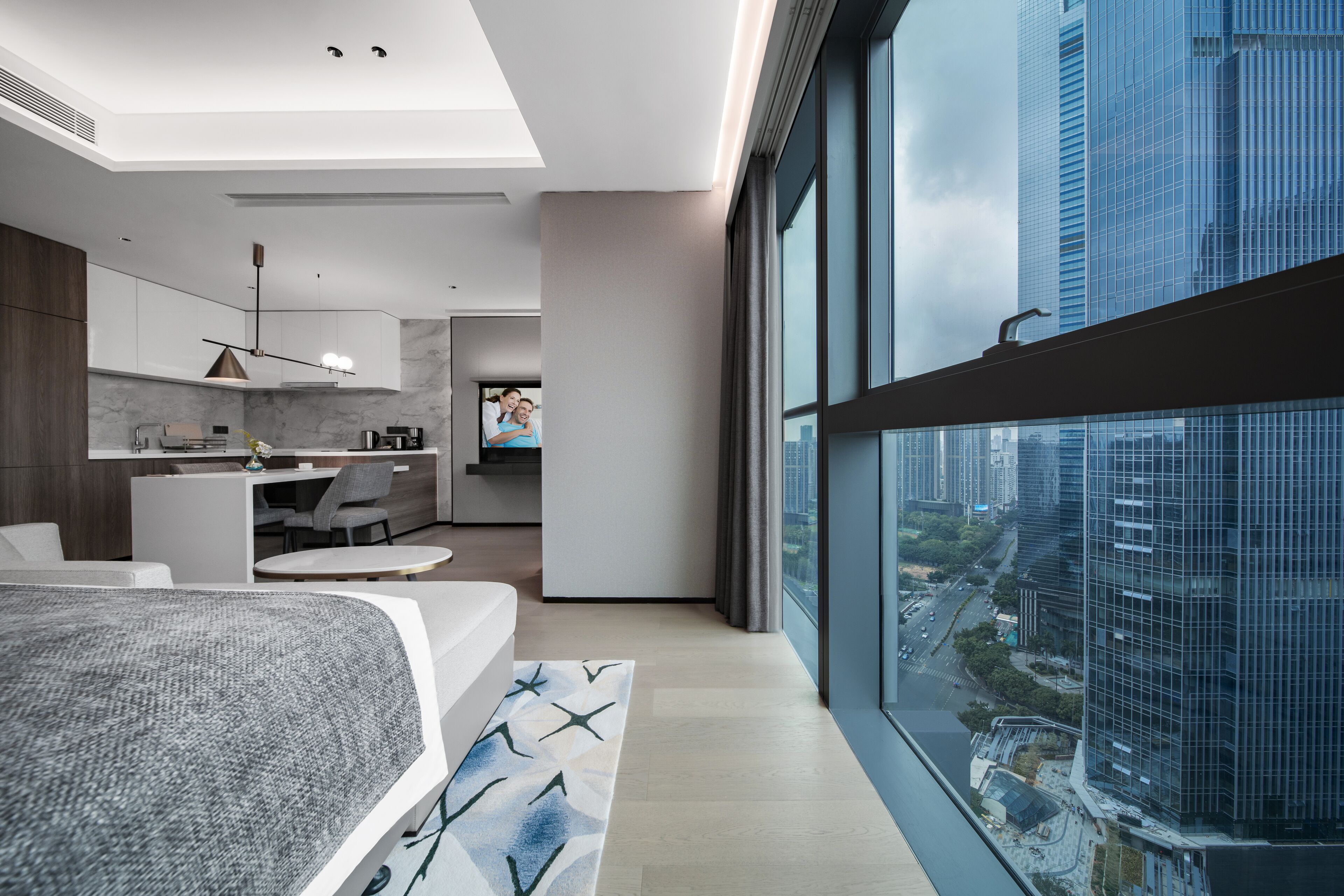 A modern bedroom with a large bed, a view of the city through floor-to-ceiling windows, a kitchen area, and a living space with a sofa and coffee table.