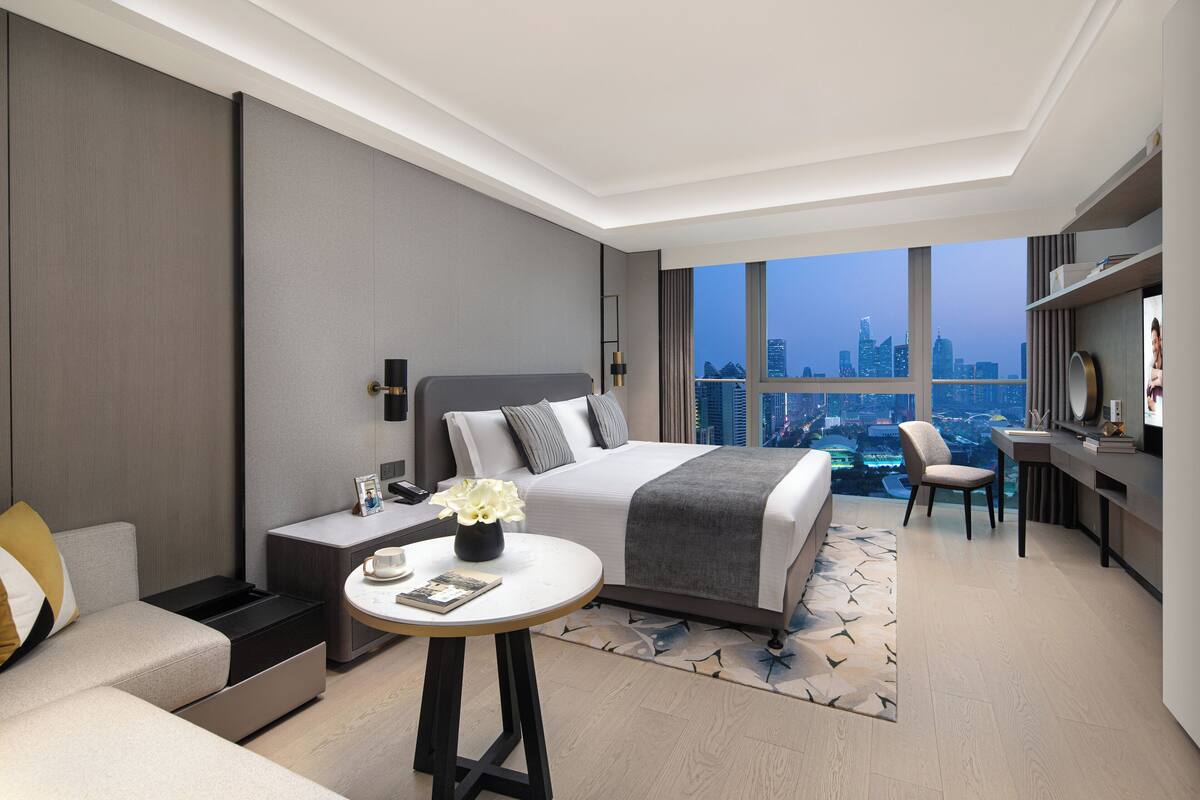 Una habitación de hotel moderna con una cama grande, un escritorio, una silla y vistas a la ciudad a través de amplios ventanales.
