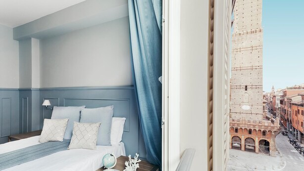 A bedroom with a bed, a nightstand, a globe, and a decorative coral piece. A window reveals a view of a historic building with arches and a tower.