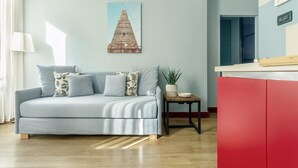A living room with a light blue sofa, a small side table with a plant, and a red kitchen area.
