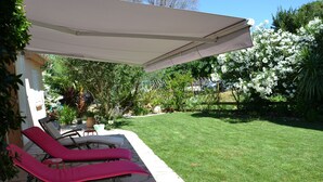 Un patio con dos sillones de descanso bajo una pérgola, un sendero de piedra y un césped cuidado con arbustos florecientes.