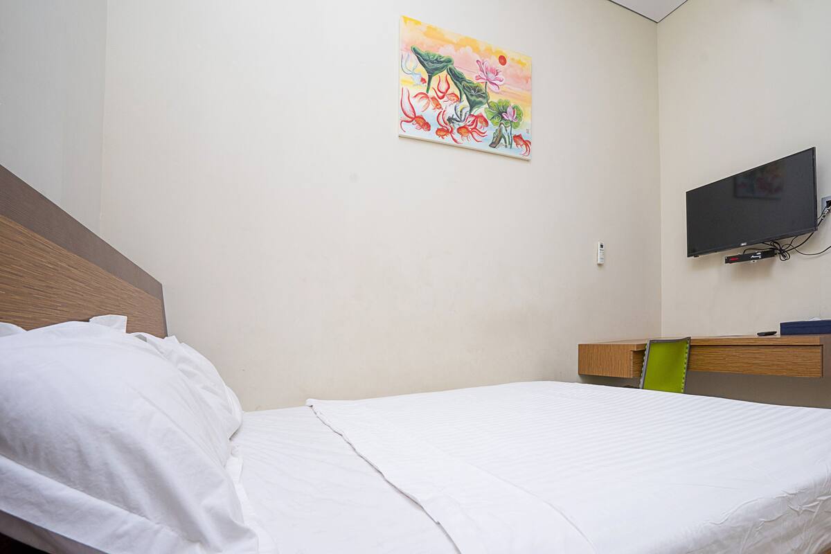 A neatly made bed with white linens, a wooden headboard, a small desk with a chair, a television mounted on the wall, and a colorful painting above the bed.