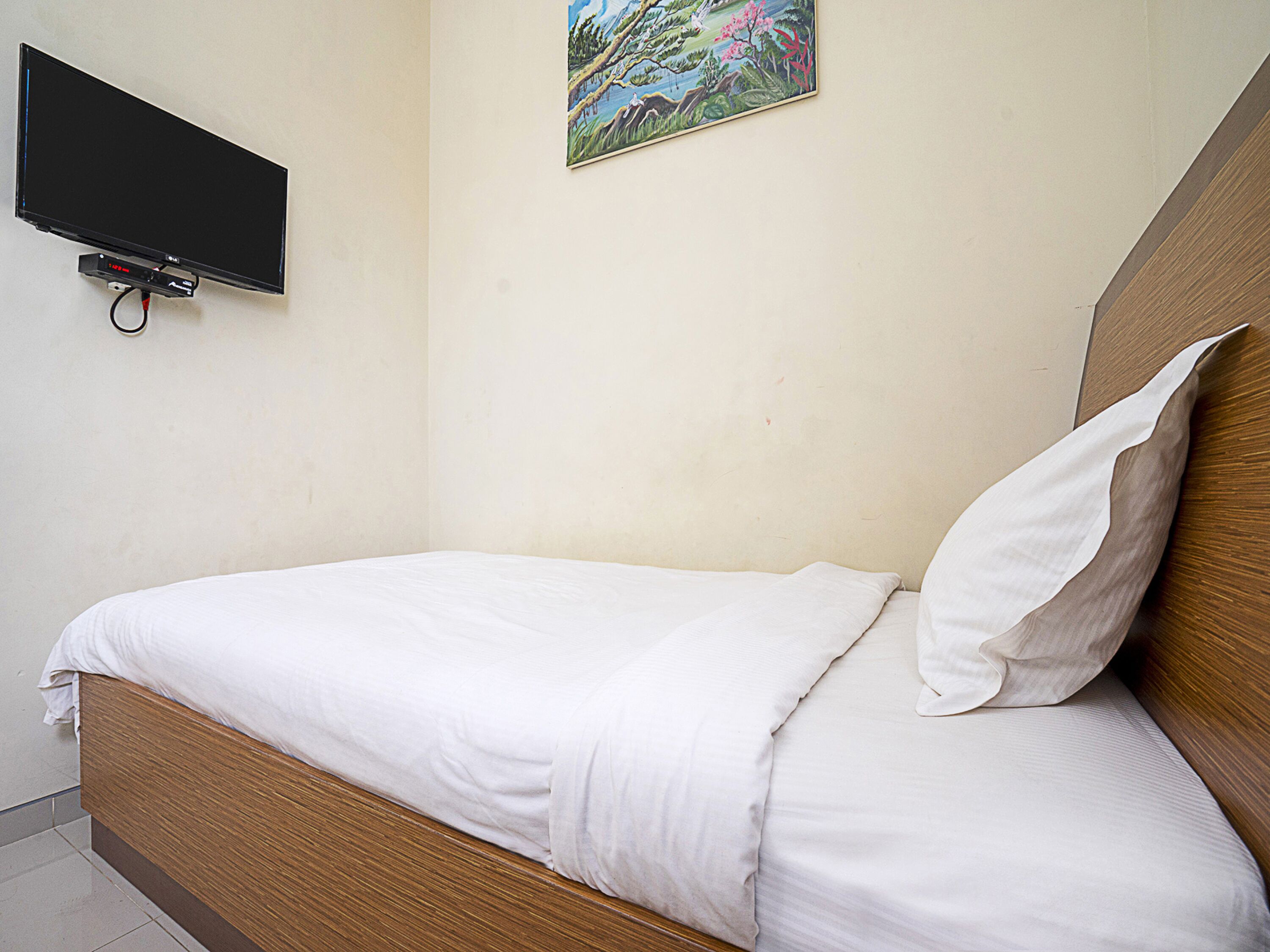 Una cama individual con ropa de cama blanca, un televisor montado en la pared y un cuadro sobre la cama.