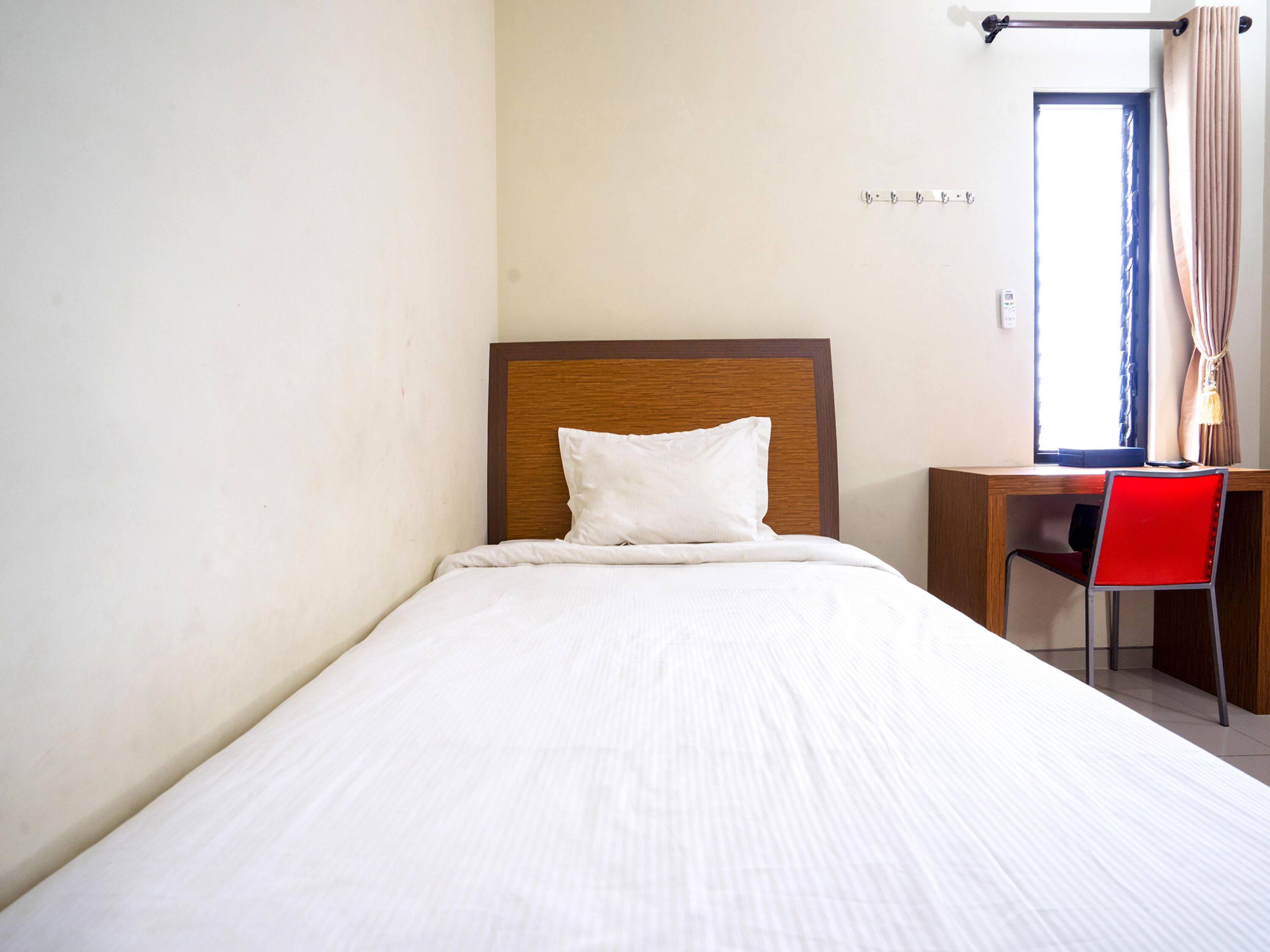 Una cama bien tendida con cabecera de madera, un escritorio con silla y una ventana con cortinas.
