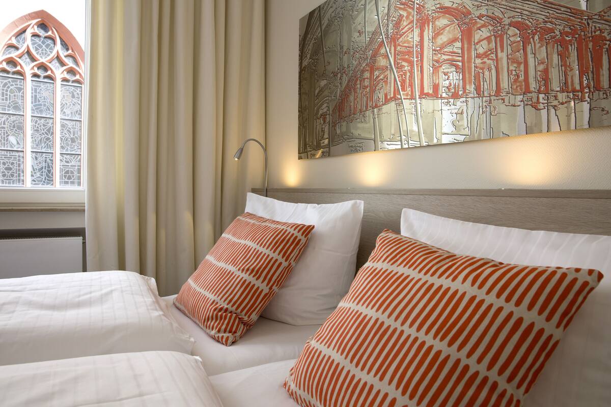 A hotel room with a large window, a bed with white bedding and orange pillows, and a wall-mounted painting.