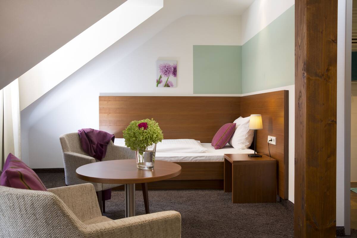 A modern hotel room with a wooden headboard, a round table, two armchairs, a nightstand, and a wall-mounted lamp.