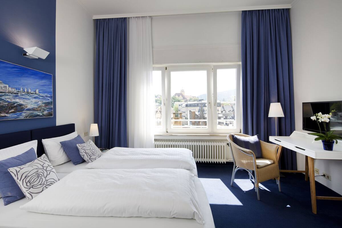 A hotel room with two beds, a desk, a chair, a TV, and a large window with curtains.